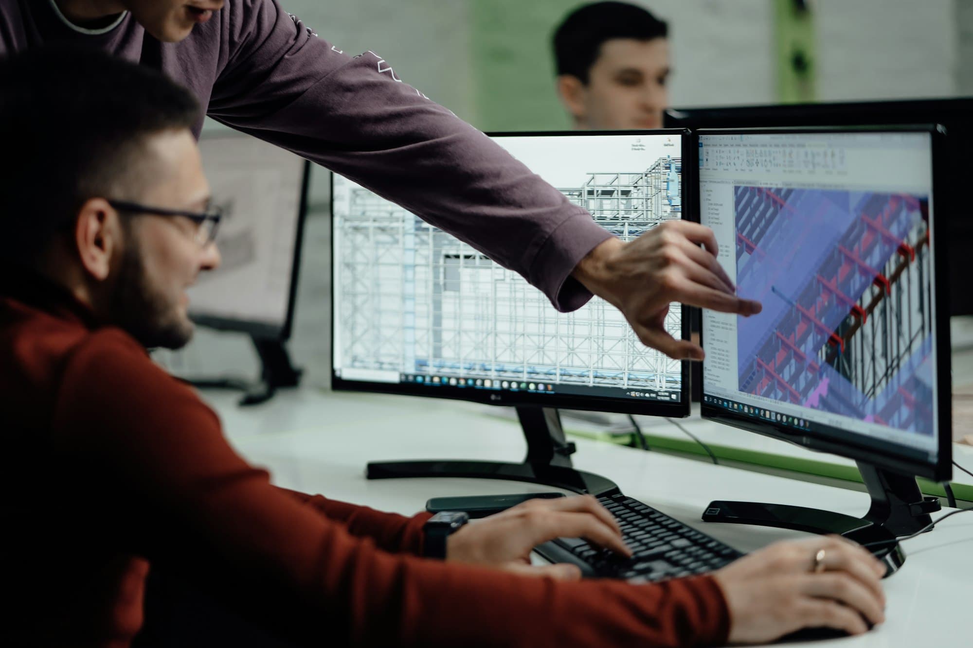 An engineering lead pointing out a detail on a 3D design model to a colleague at dual monitors